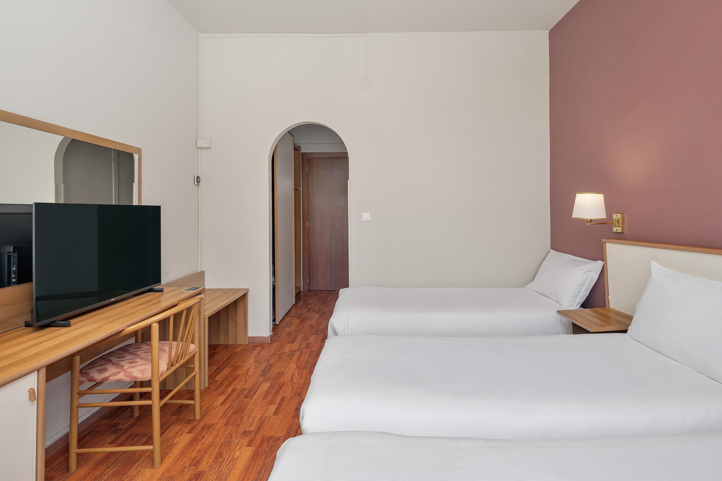 A hotel room with a TV, a desk, two beds, and a hallway.