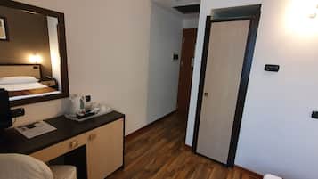 Habitación de hotel con cama, mesita de noche y espejo. El suelo es de madera y se ve una puerta al fondo.
