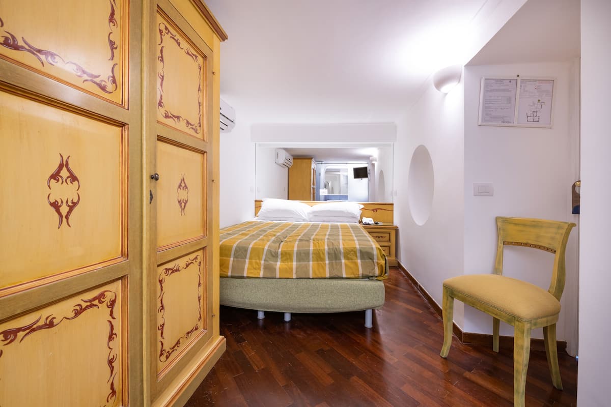 A bedroom with a bed, a wooden wardrobe, a chair, and a nightstand.