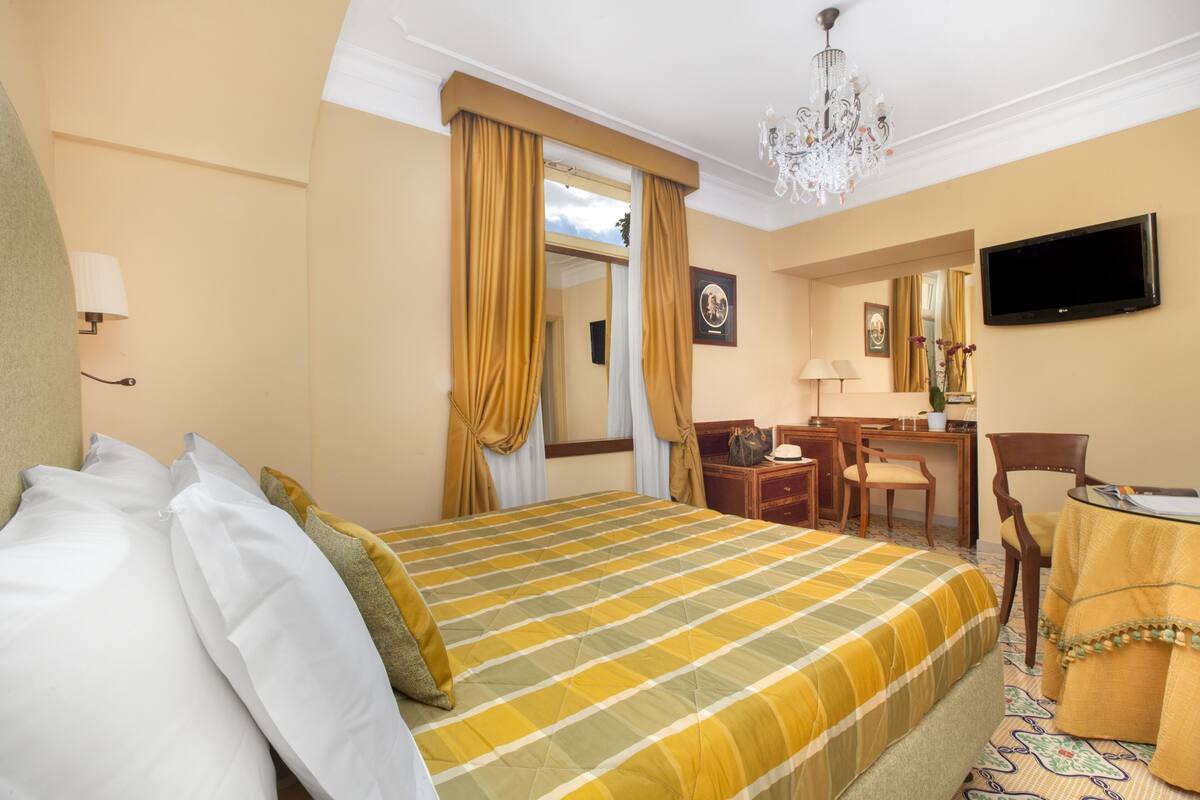A hotel room with a large bed, a desk with a chair, a television, and a chandelier.
