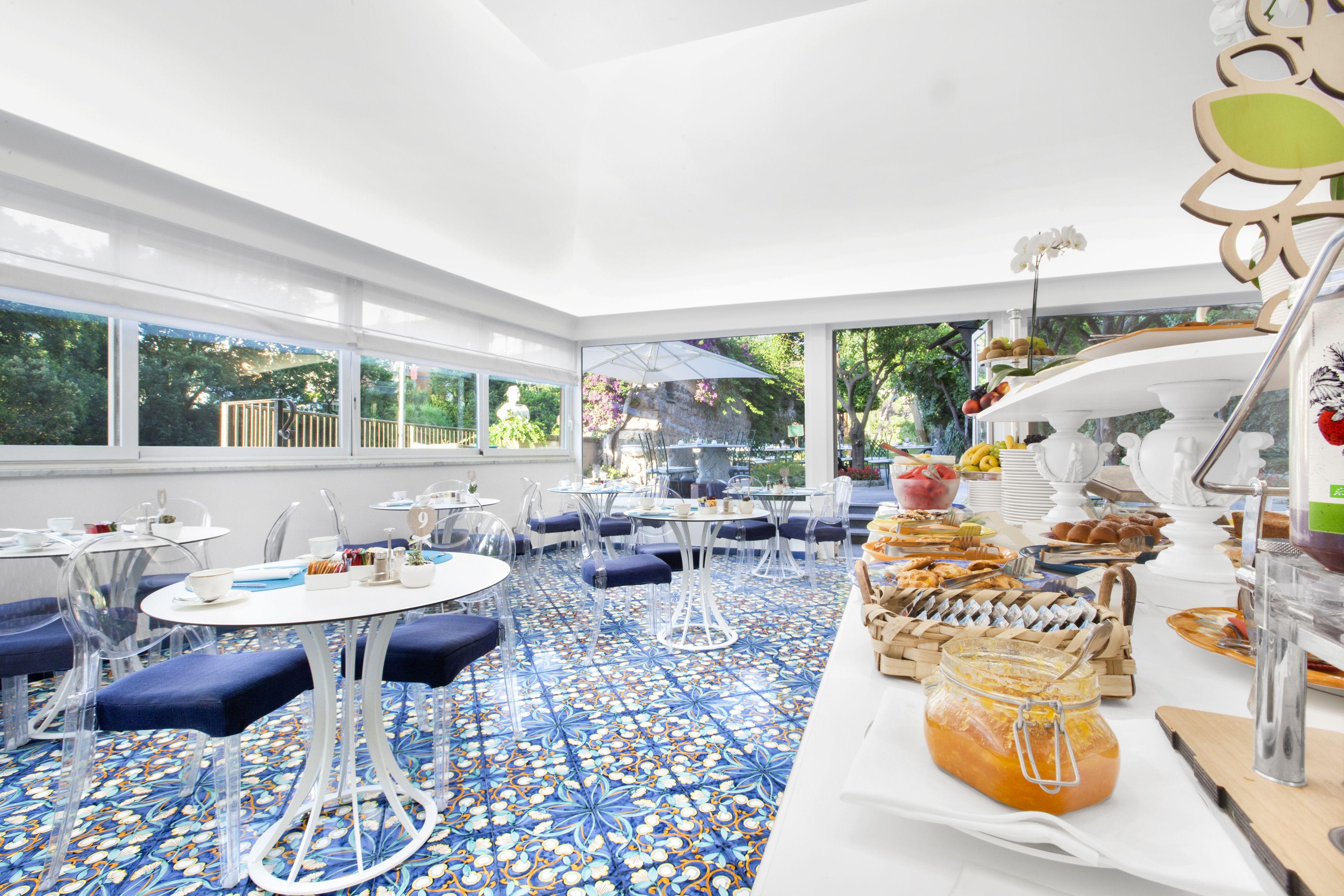 A bright, open dining area with round tables and blue cushioned chairs, a tiled floor, and a buffet with various food items.