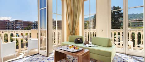 A balcony with a green sofa, a wooden table with a tray of fruit and cups, and patterned floor tiles.
