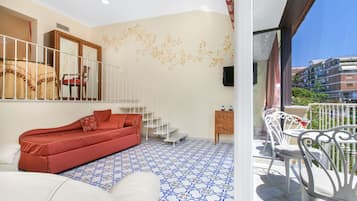A bedroom with a red sofa, a white bed, and a patterned floor. There is a balcony with white chairs and a view of buildings.