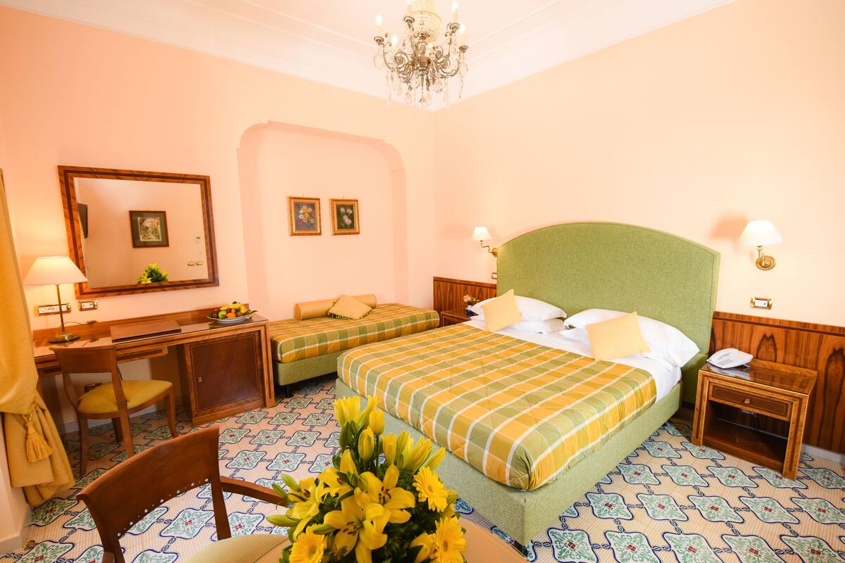 A hotel room with two beds, a desk, a chair, a chandelier, and a mirror.