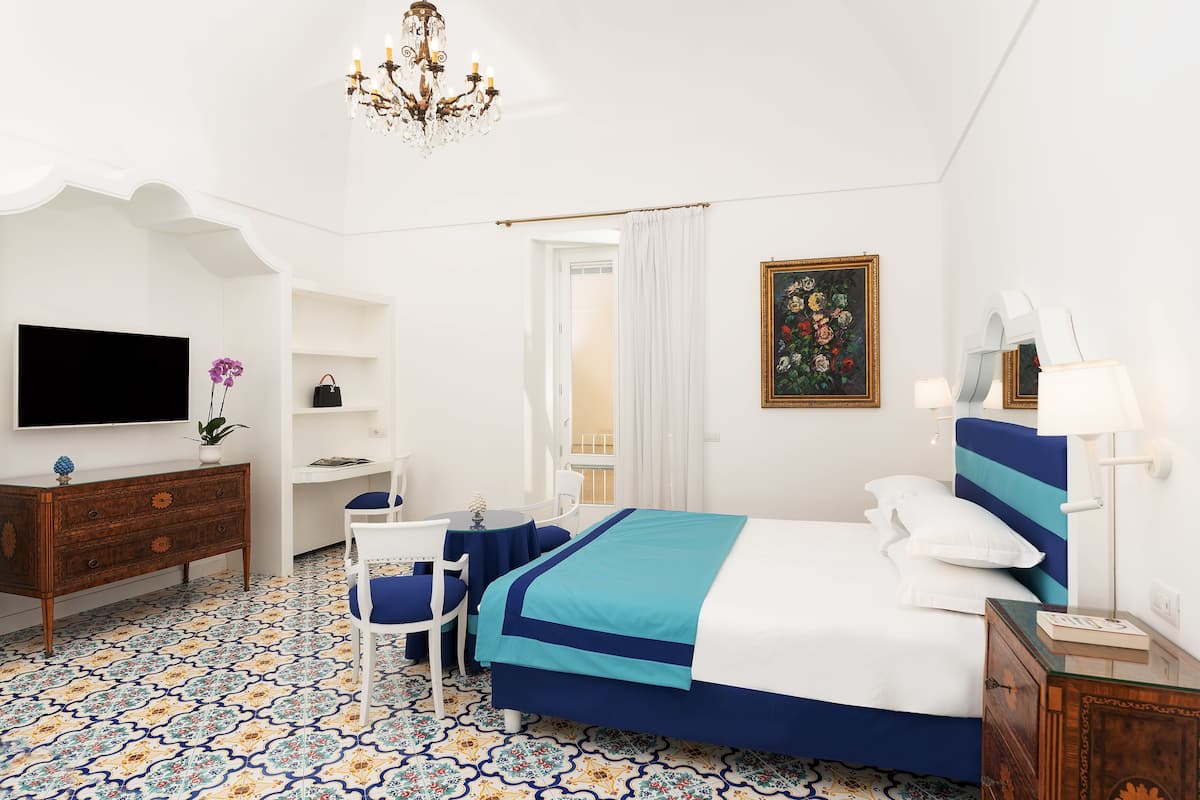 A bedroom with a bed, a desk, a chair, a TV, and a chandelier.