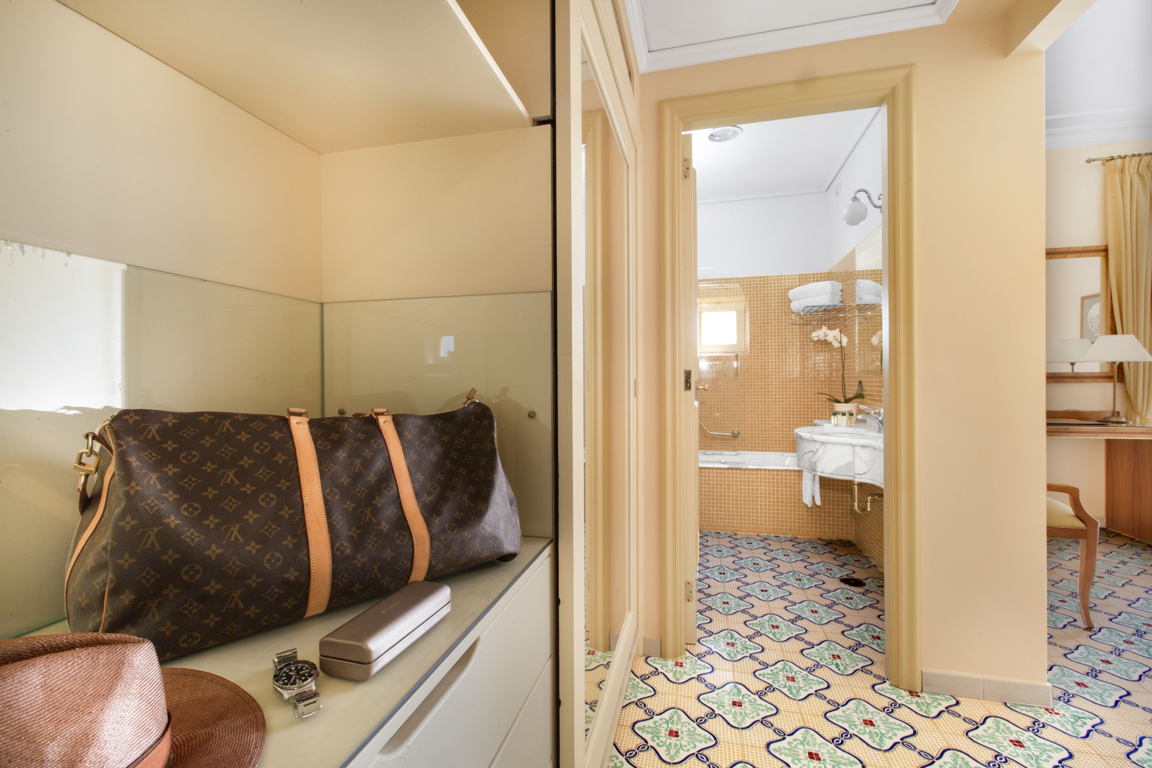 A hotel room with a bed, a suitcase, a watch, and a desk with a lamp.