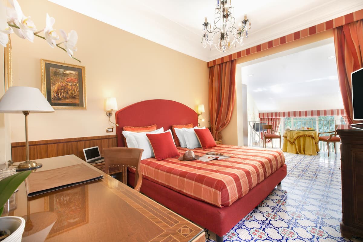 A hotel room with a large bed, a desk with a laptop, a chair, a table with a lamp, and a view of the outside.