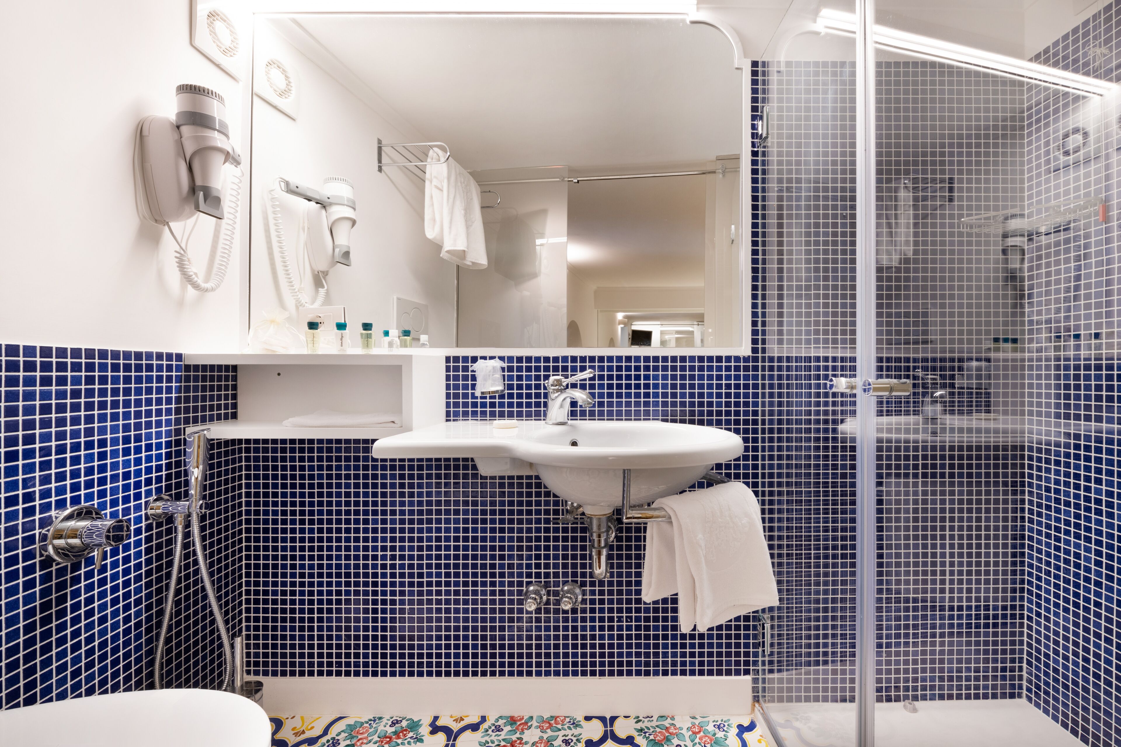 A bathroom with a white sink, blue tiled walls, a glass shower enclosure, and a hairdryer mounted on the wall.