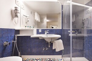 Un baño con un lavamanos blanco, paredes con azulejos azules, una mampara de cristal para la ducha y un secador de pelo fijado en la pared.
