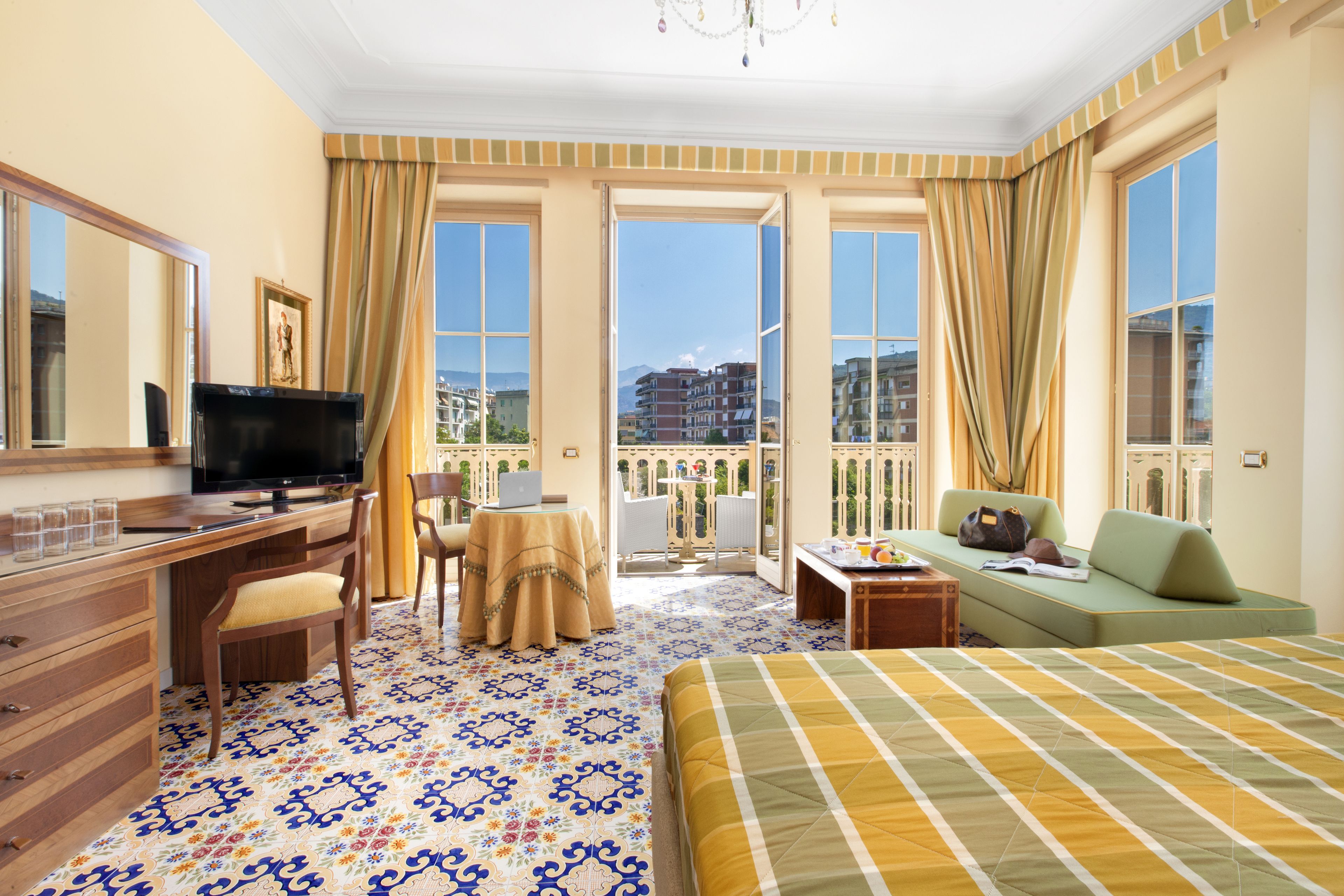 A hotel room with a bed, a desk with a computer, a TV, and a balcony with a view of buildings.