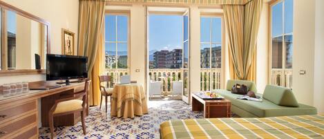 A hotel room with a bed, a desk with a computer, a TV, and a balcony with a view of buildings.