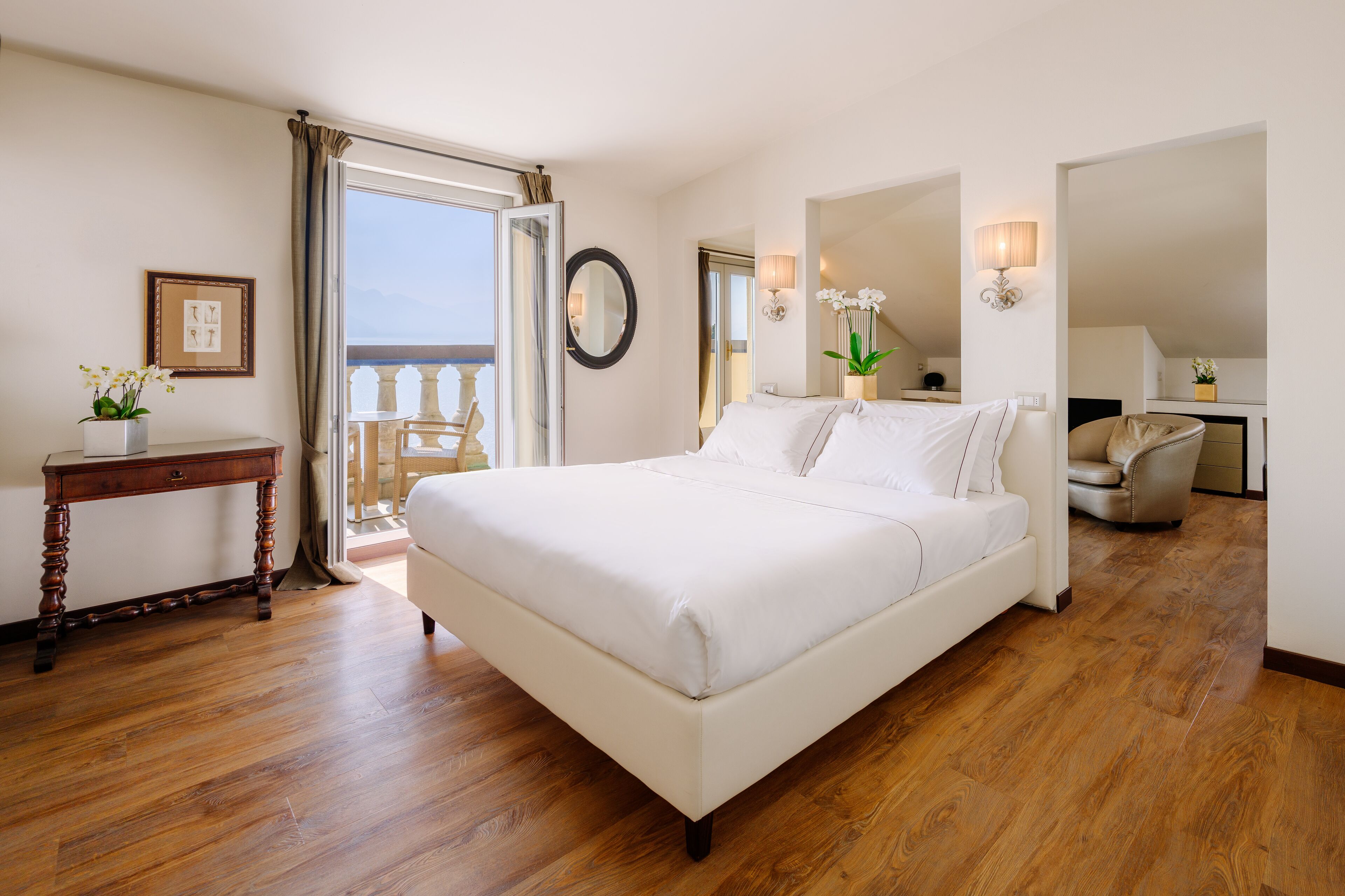 A bedroom with a large bed, wooden flooring, a desk, and a balcony with a view.