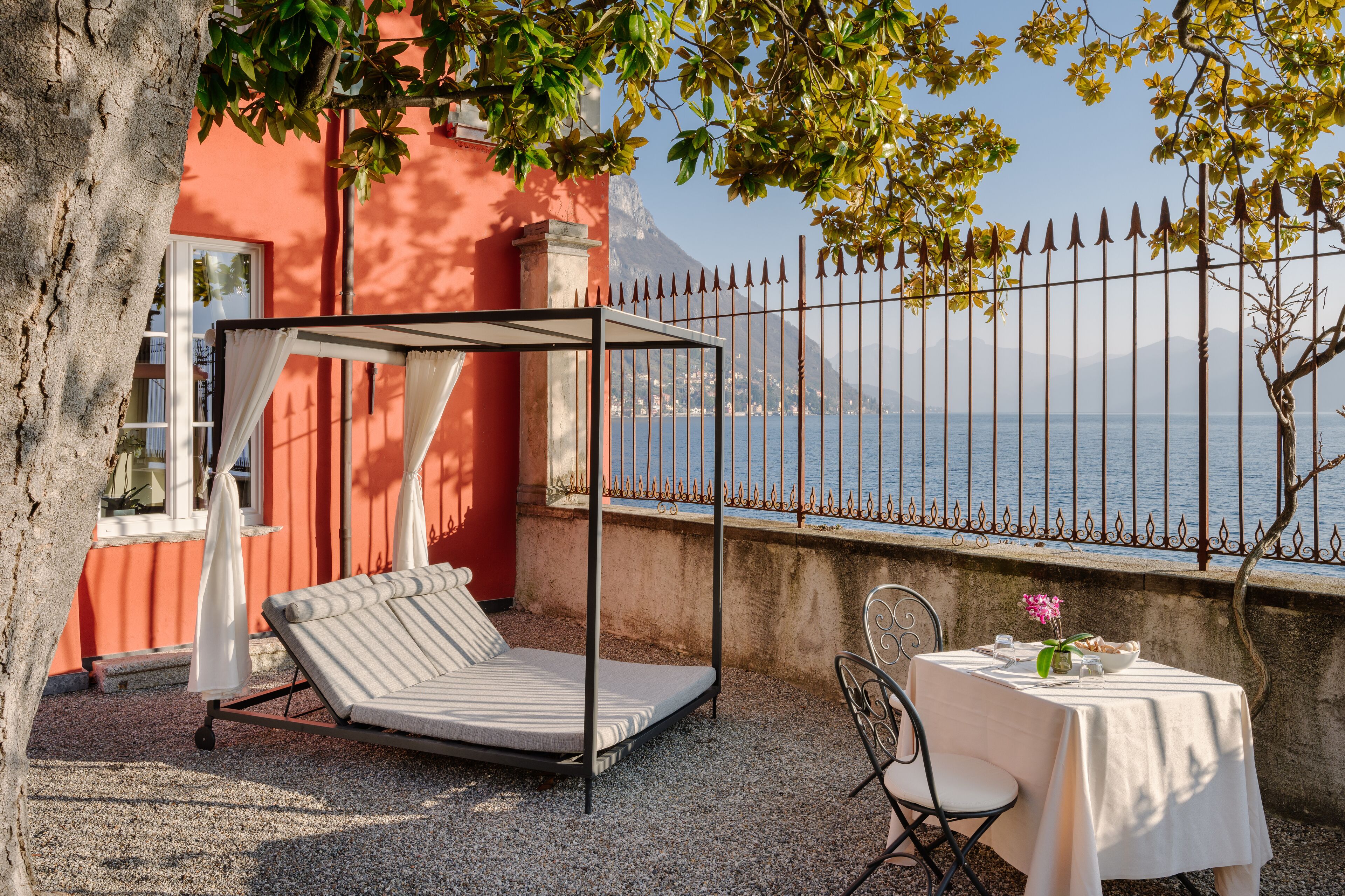 Un transat sous un auvent, une table avec un vase de fleurs et une vue sur la mer à travers une clôture en métal.