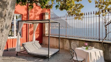 A sunbed under a canopy, a table with a vase of flowers, and a view of the sea through a metal fence.