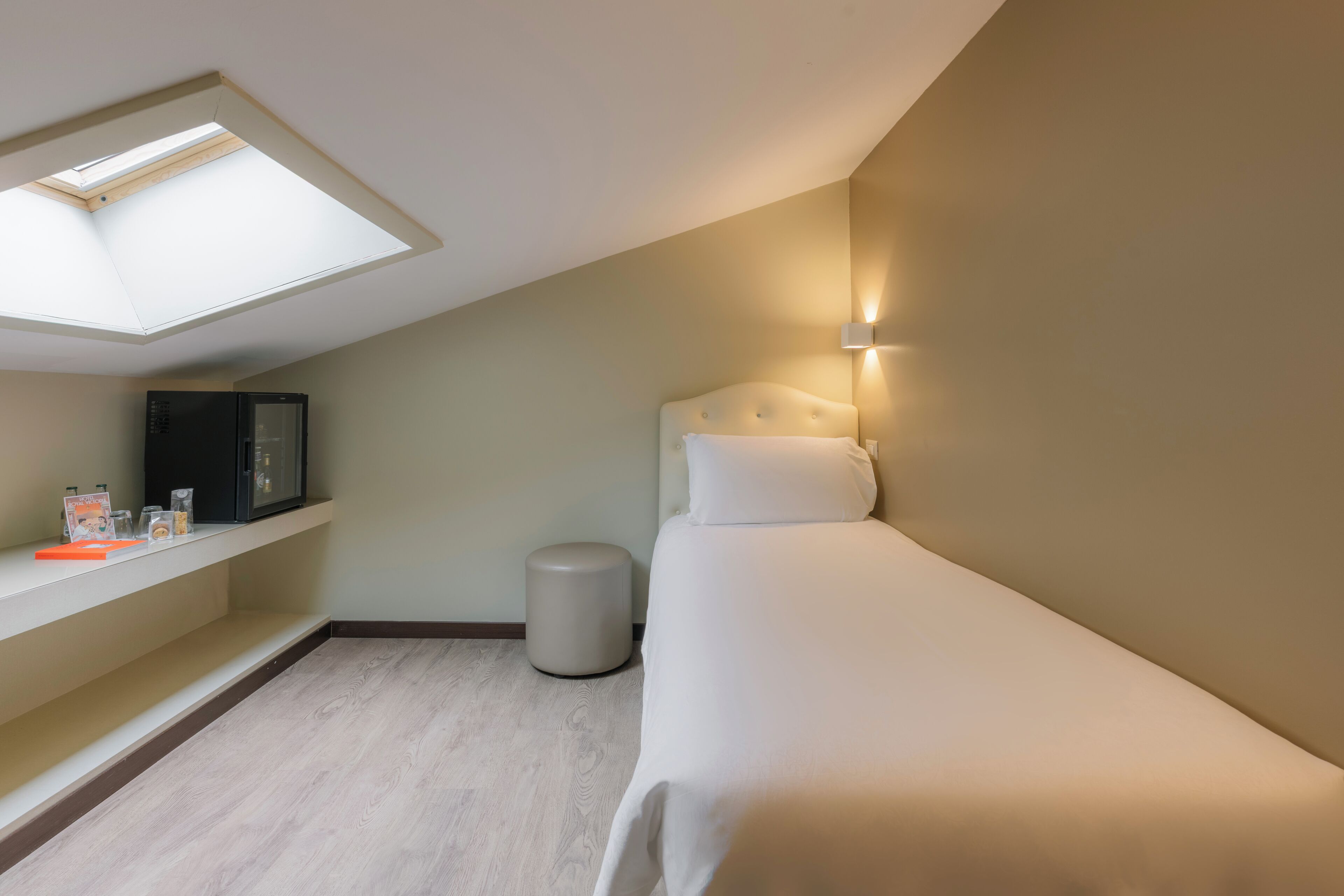 Ein Schlafzimmer mit Oberlicht, einem Bett, einem Nachttisch, einem kleinen Hocker und einer Wandleuchte.