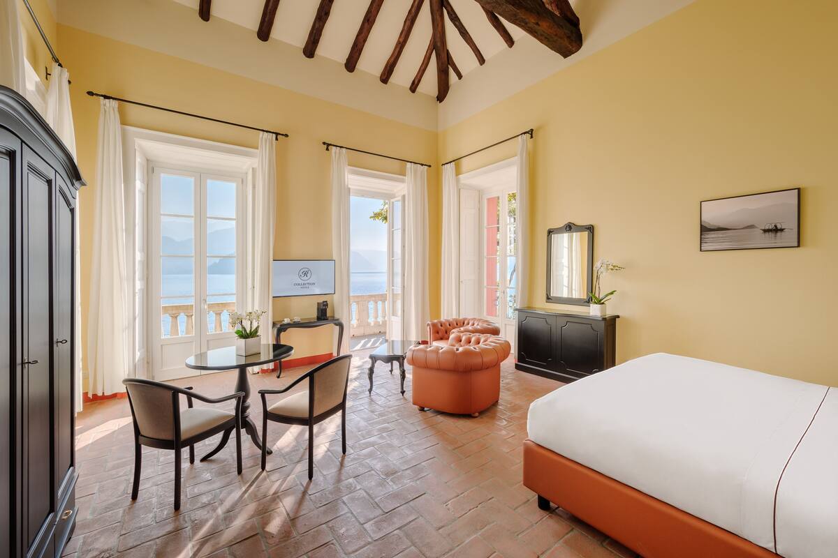 A room with a bed, a dining table with chairs, a small table with a lamp, and a view of the sea through large windows.