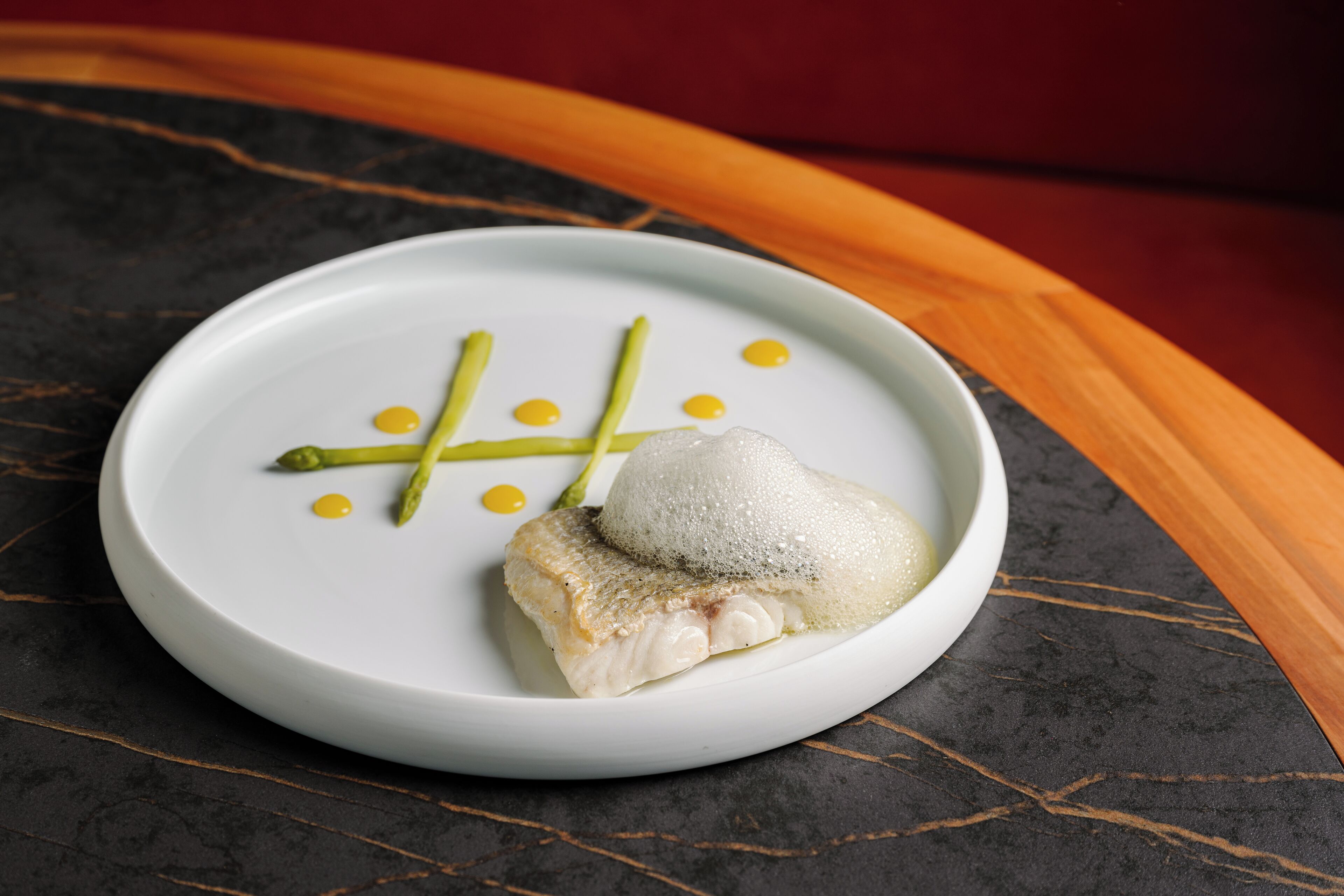 Um prato branco com um pedaço de peixe, aspargos e pontos amarelos sobre uma superfície de mármore escuro.