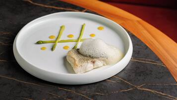 Um prato branco com um pedaço de peixe, aspargos e pontos amarelos sobre uma superfície de mármore escuro.