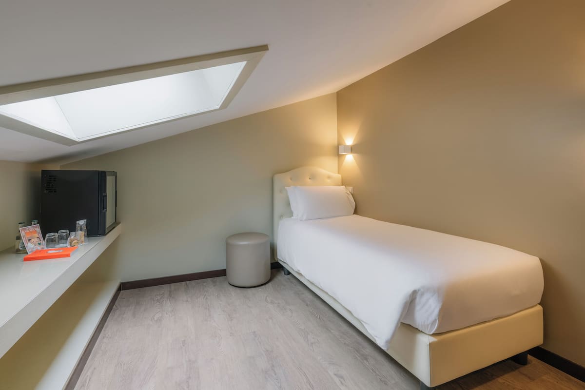 A bedroom with a skylight, a bed, a bedside table with a lamp, and a small trash bin.