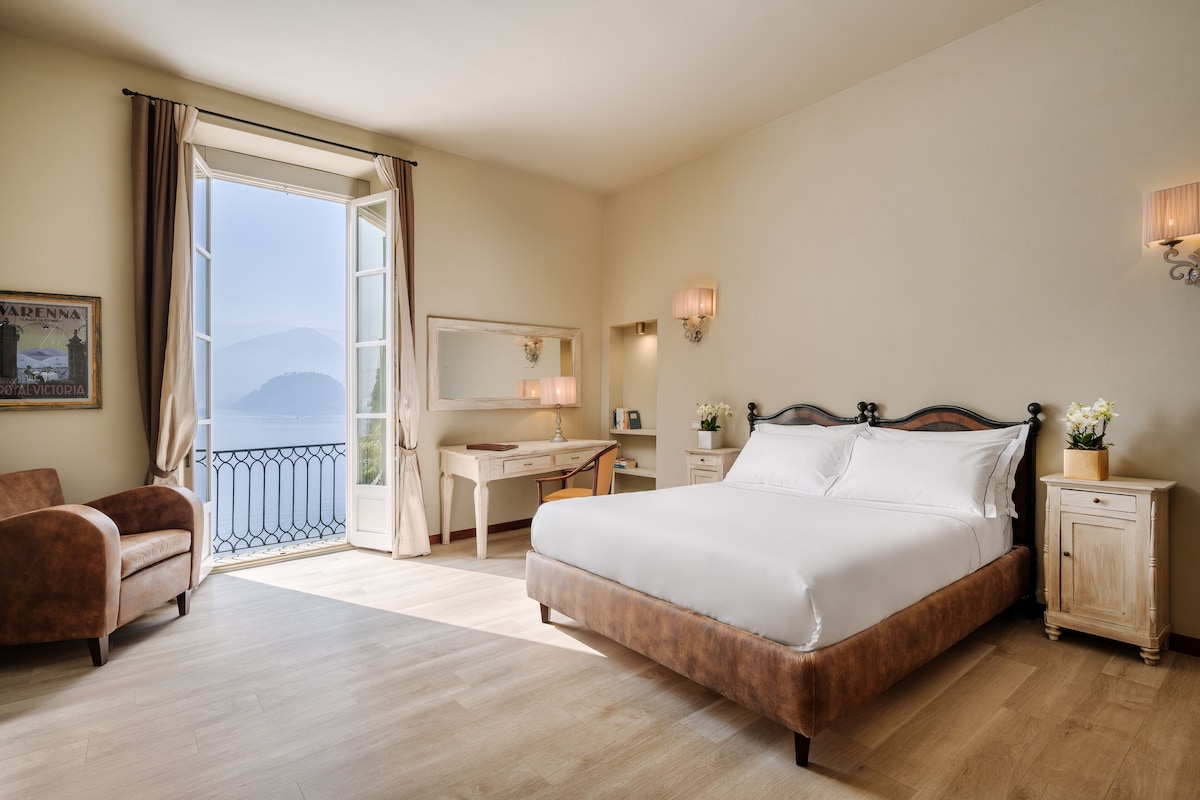 A hotel room with a large bed, a desk, a chair, a nightstand, a mirror, and a view of the outdoors.