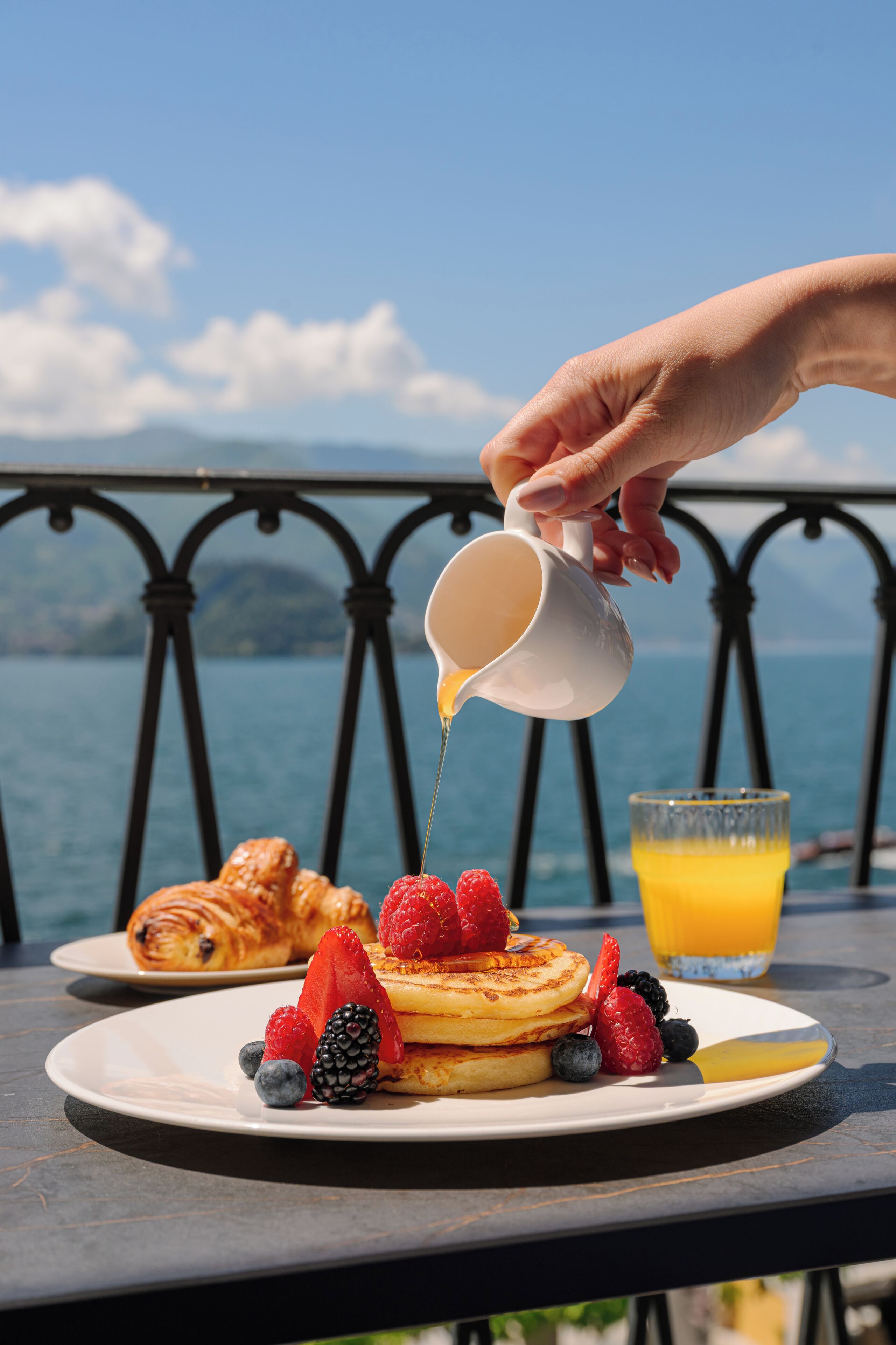 Une table dressée pour le petit-déjeuner, avec des crêpes, des fruits rouges et un croissant, le tout placé devant un paysage pittoresque.