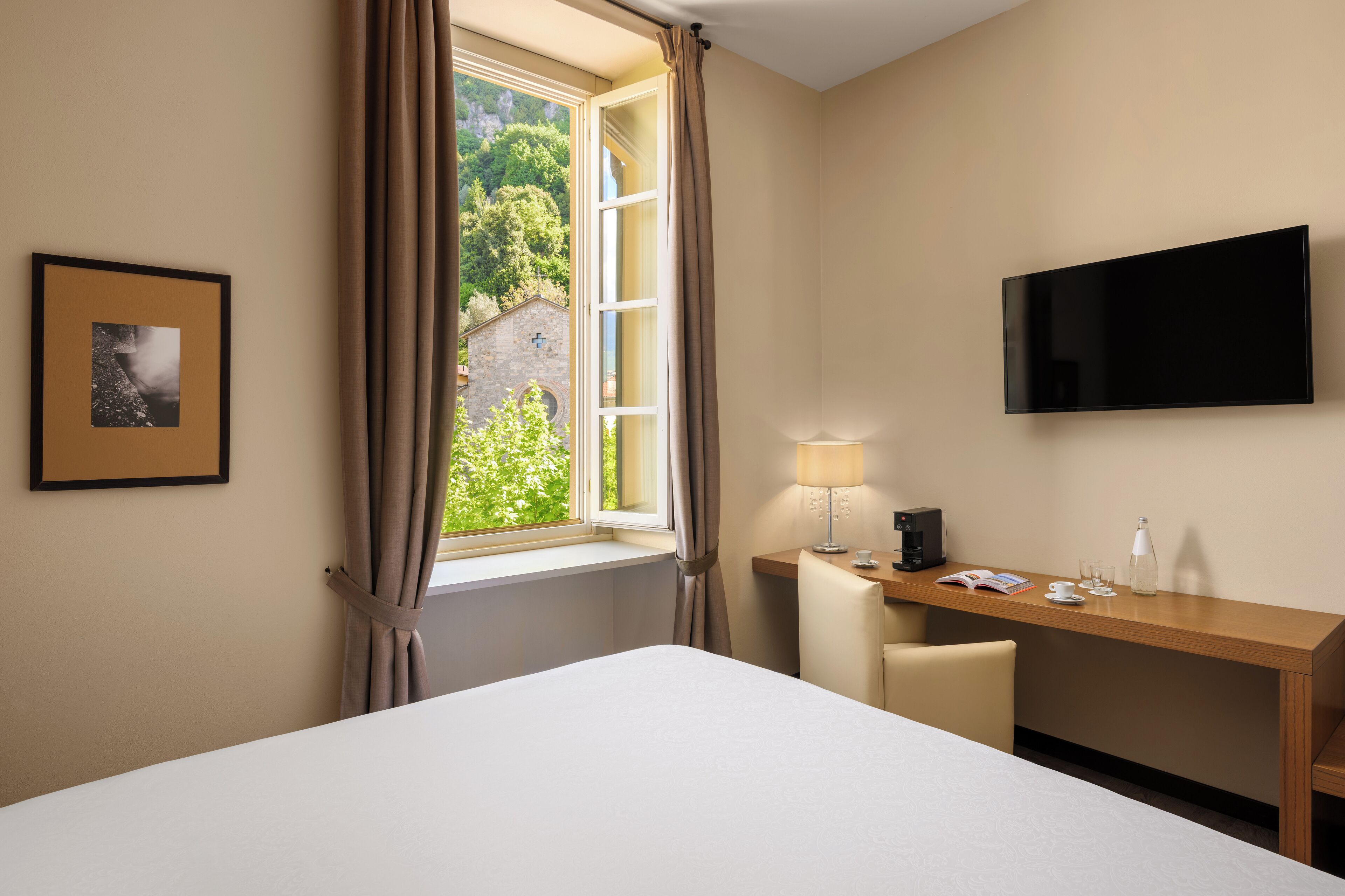 Une chambre d’hôtel avec un lit, un bureau, une télévision, une fenêtre avec des rideaux et un tableau encadré au mur.