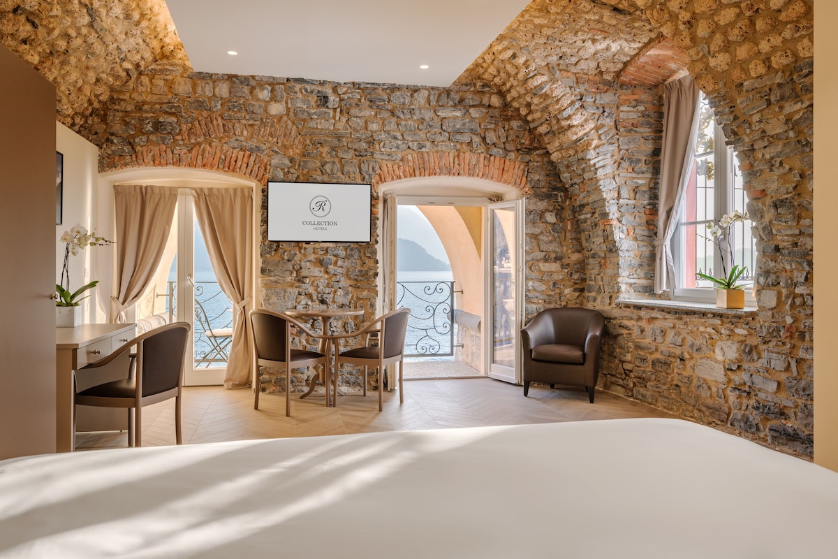 A room with stone walls, a bed, a desk with chairs, a table, and a view of the sea.