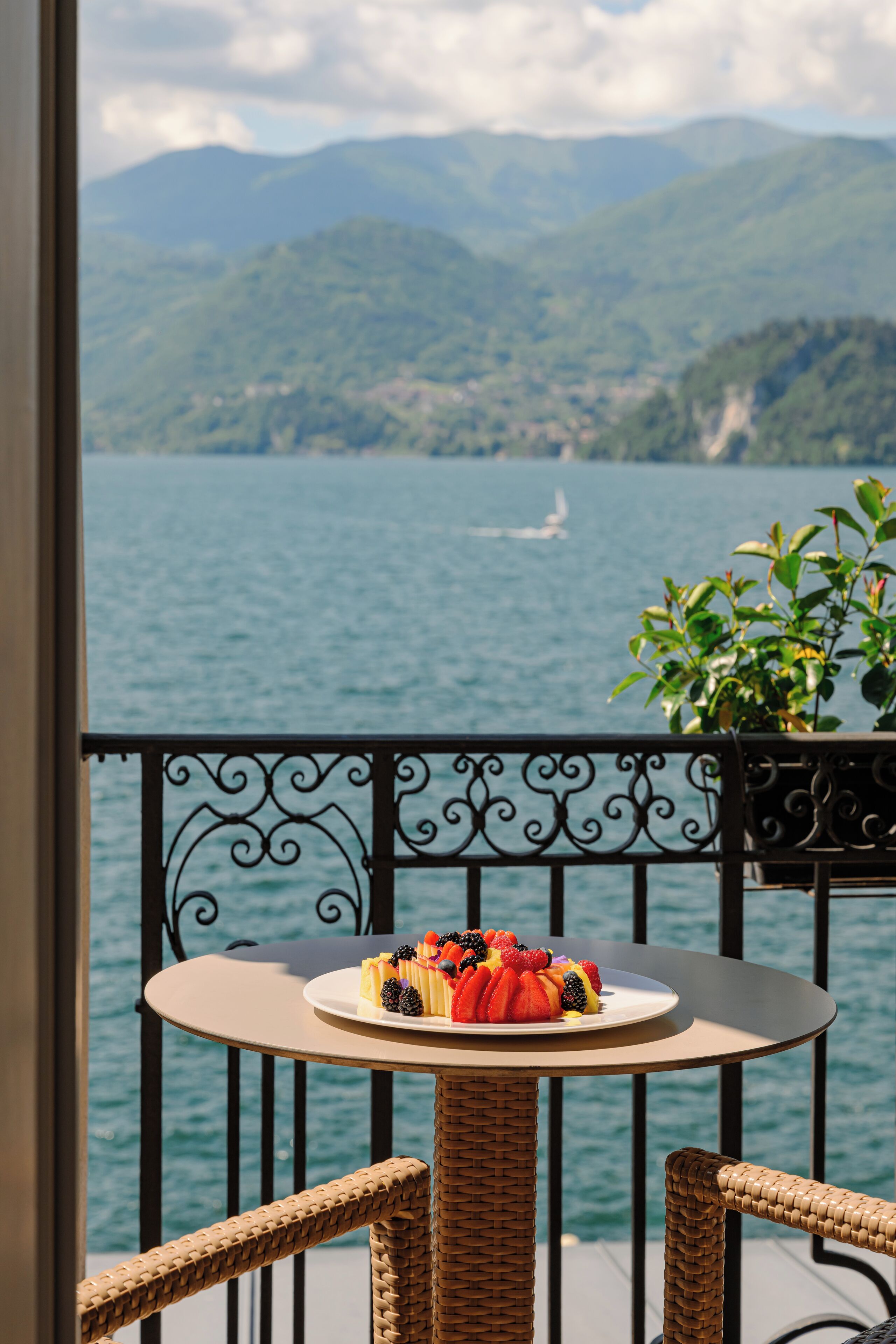 Un balcón con una mesa y sillas de mimbre, un plato de fruta y vistas a un lago con montañas al fondo.