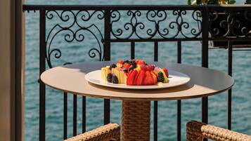 Un balcón con una mesa y sillas de mimbre, un plato de fruta y vistas a un lago con montañas al fondo.
