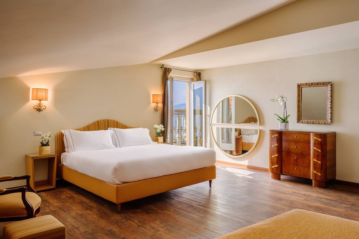 A hotel room with a large bed, a wooden dresser, a round mirror, and a view of the outdoors.