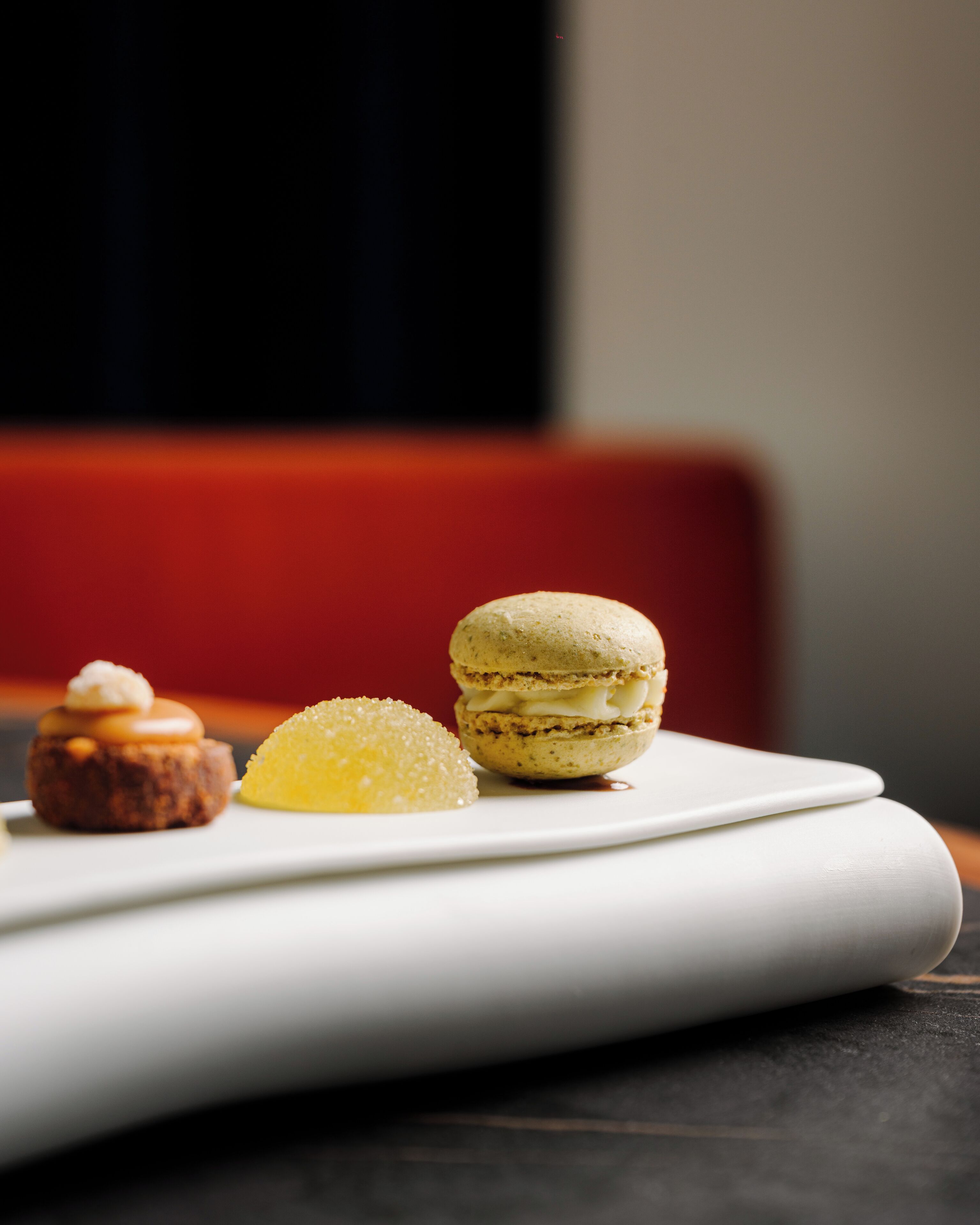 Ein weißer Teller mit drei Desserts: einem Macaron, einem kleinen Törtchen und einem Zitronenschnitzel.