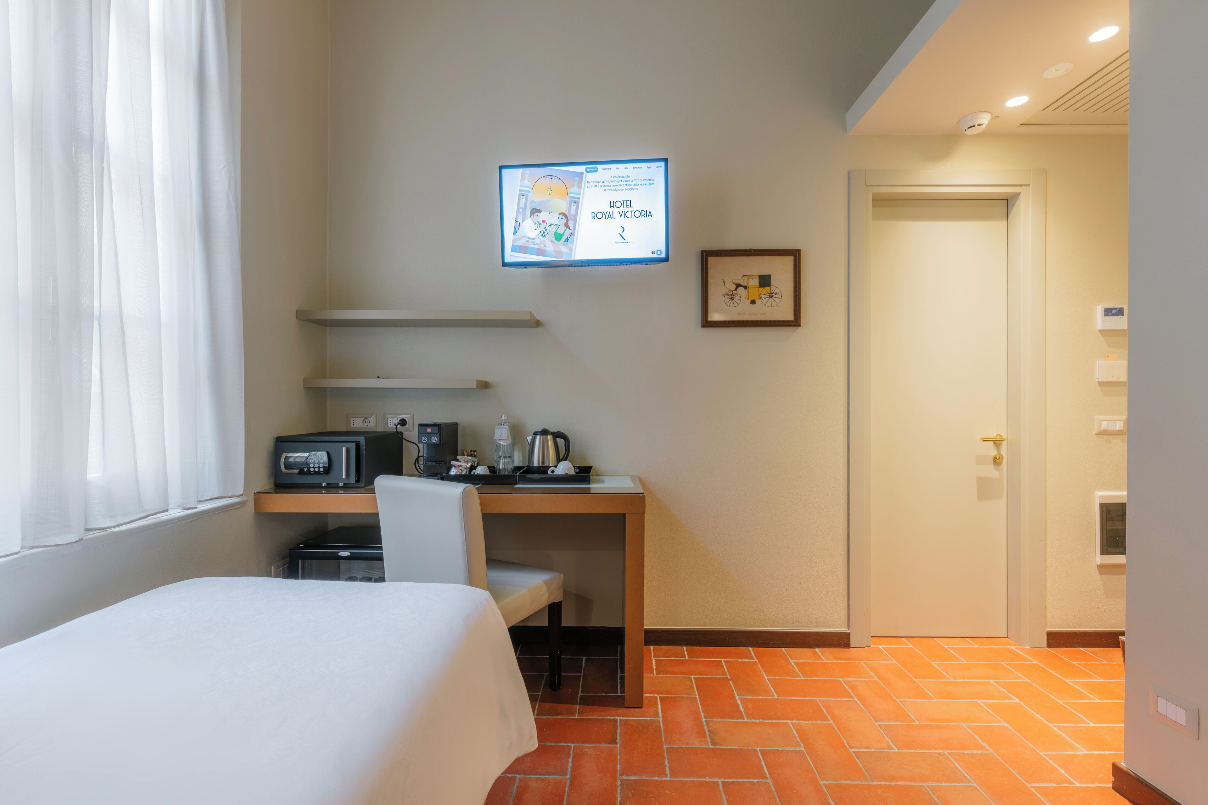Habitación de hotel con cama, escritorio con computadora, cafetera y televisión mostrando el nombre del hotel.