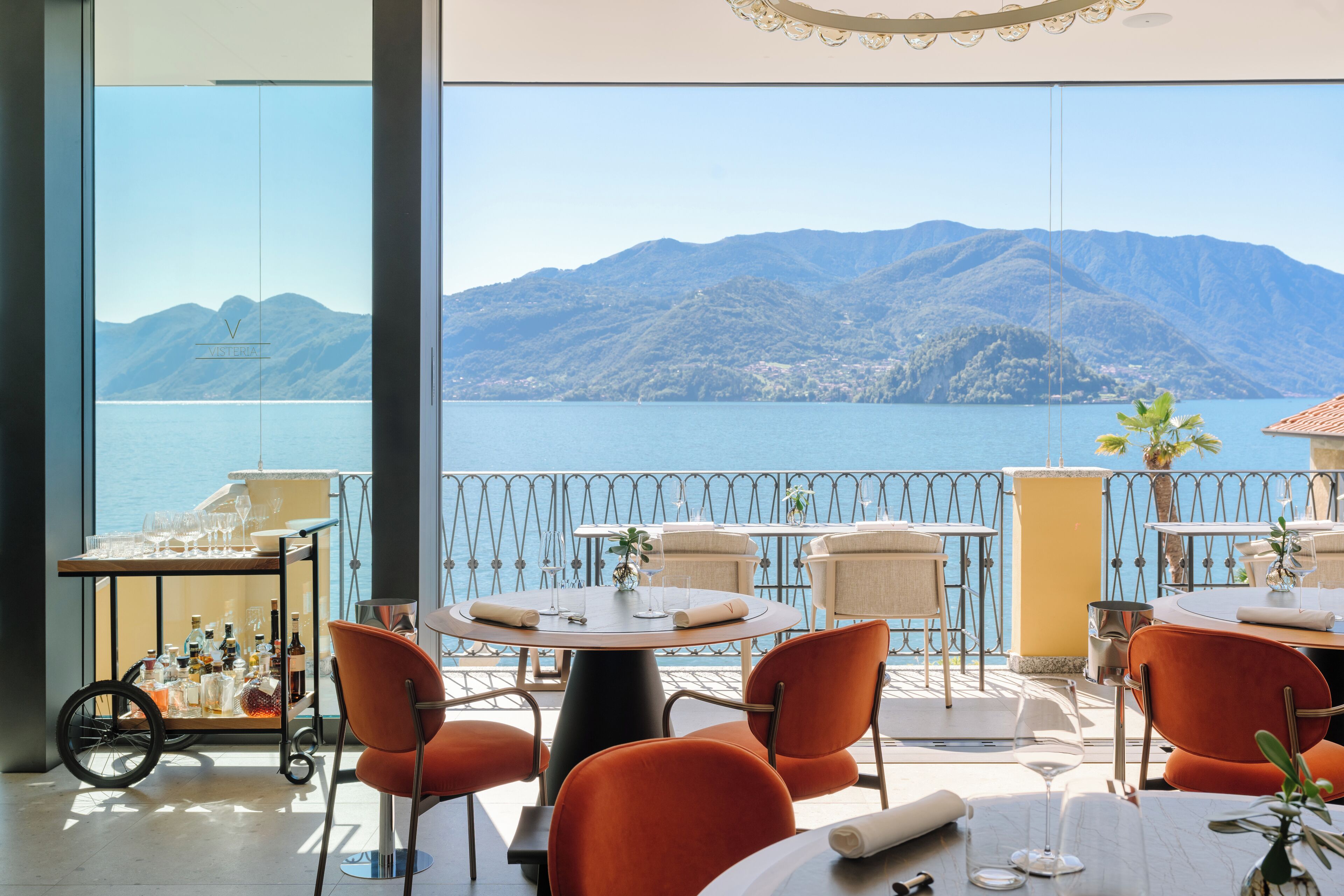 Una terraza con mesas redondas y sillas naranjas, con vistas a un lago y montañas.