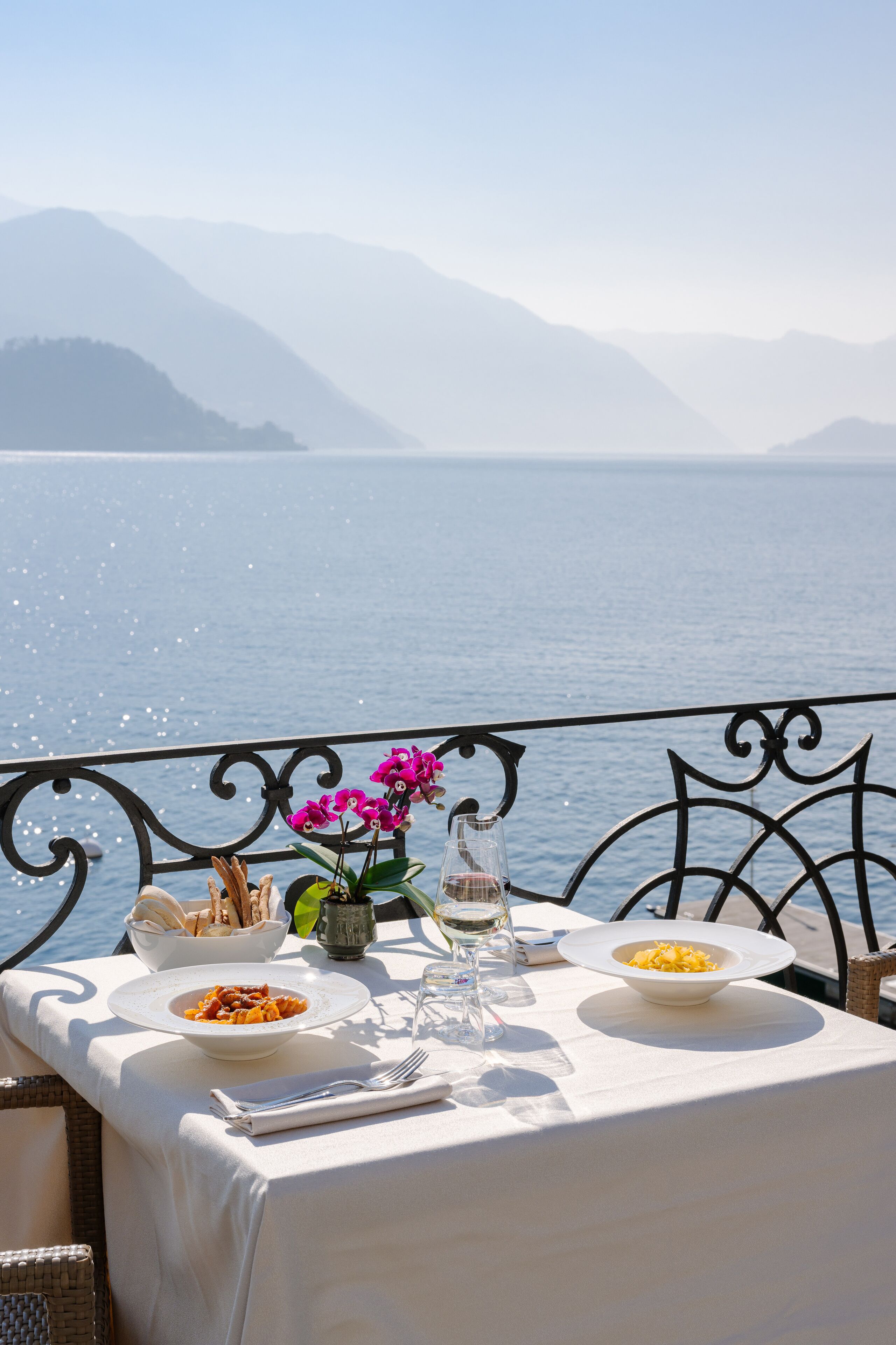 A table set with plates of food, a glass of water, and a vase of flowers, overlooking a body of water with mountains in the background.