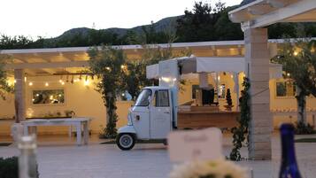 A white vintage car parked in a courtyard with a table and chairs, surrounded by greenery and a building with string lights.