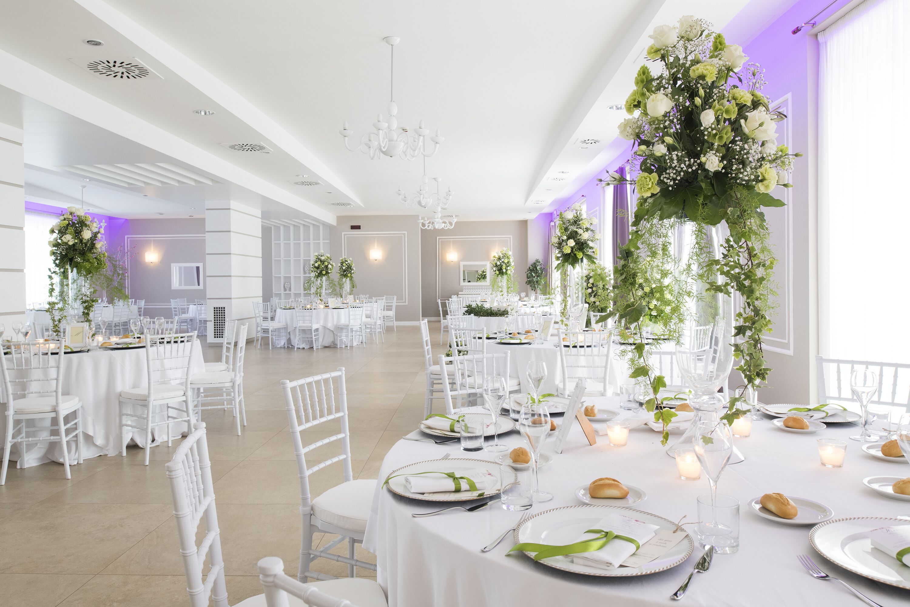 A spacious banquet hall with white tables and chairs, floral centerpieces, and candle lighting.
