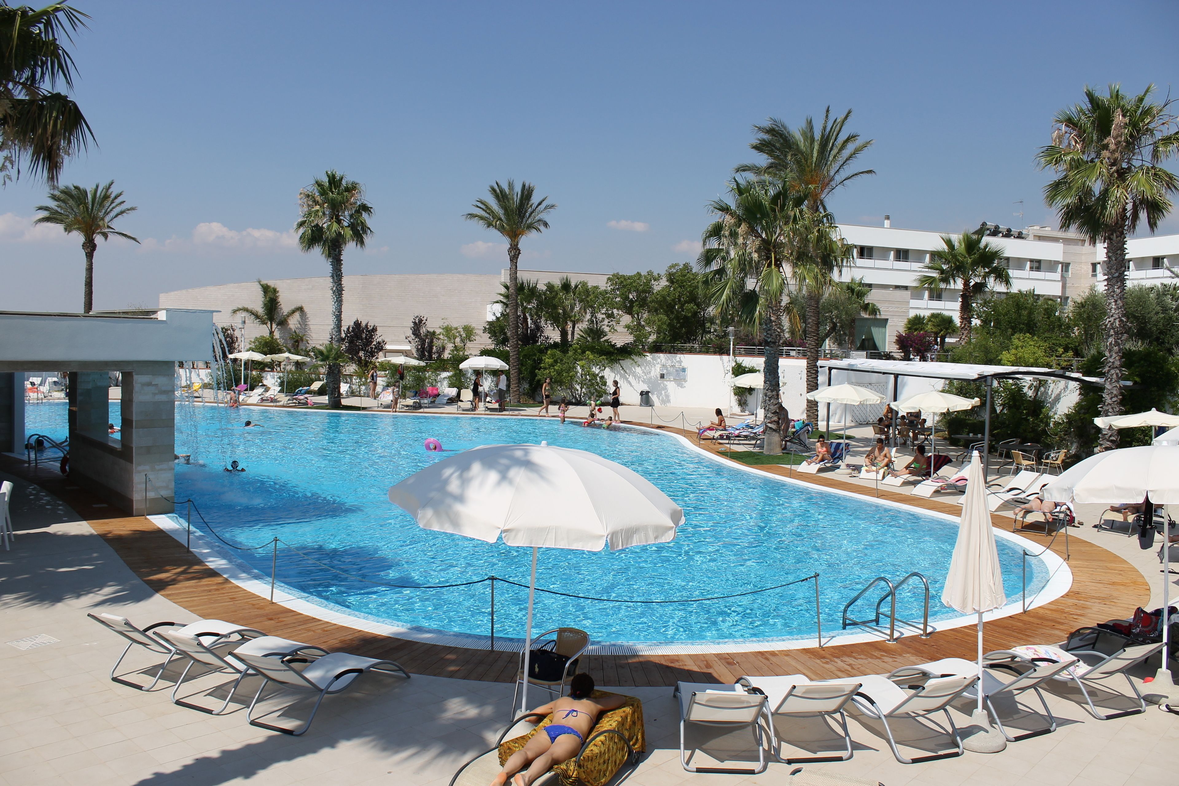 A swimming pool with sun loungers and umbrellas, surrounded by palm trees and a building in the background.
