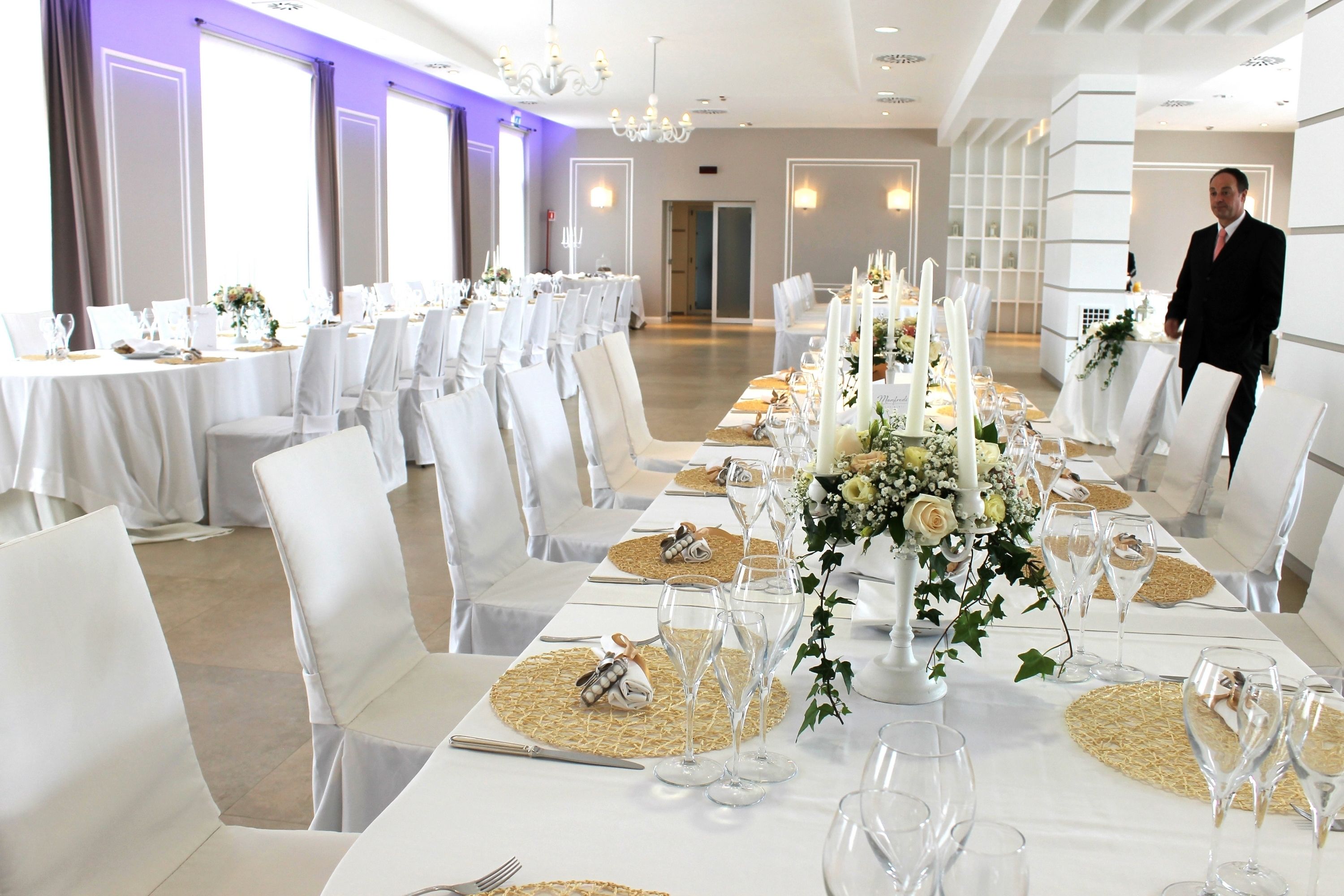 A banquet hall with long tables set for a formal event, featuring white tablecloths, gold placemats, and candle centerpieces.