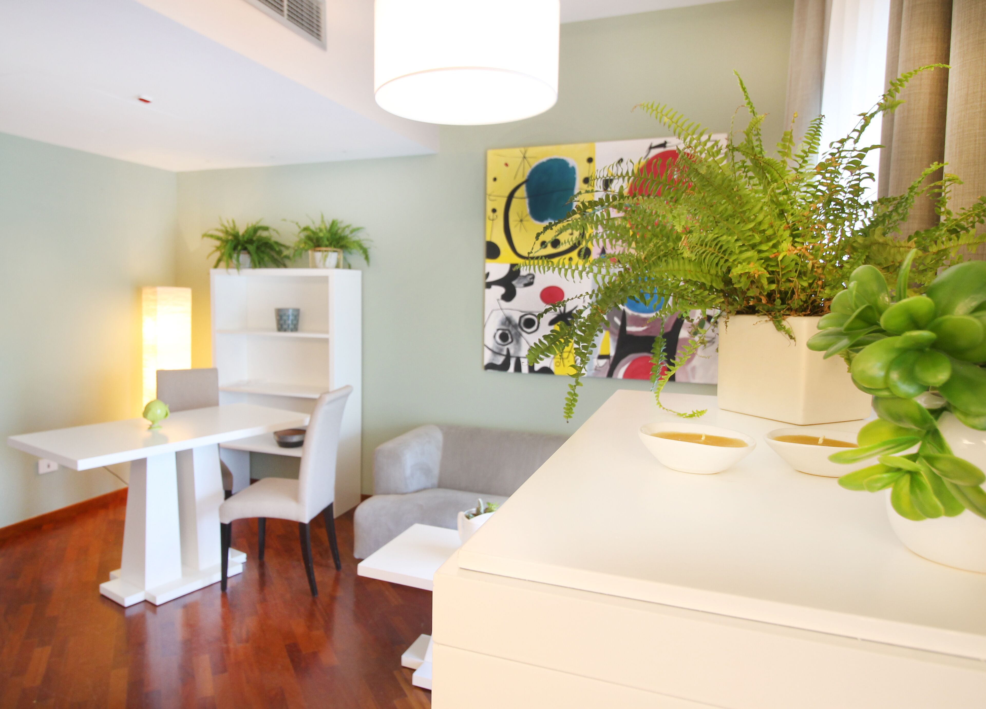 A modern living room with a white sofa, a dining table, and a white cabinet. There are potted plants and a painting on the wall.