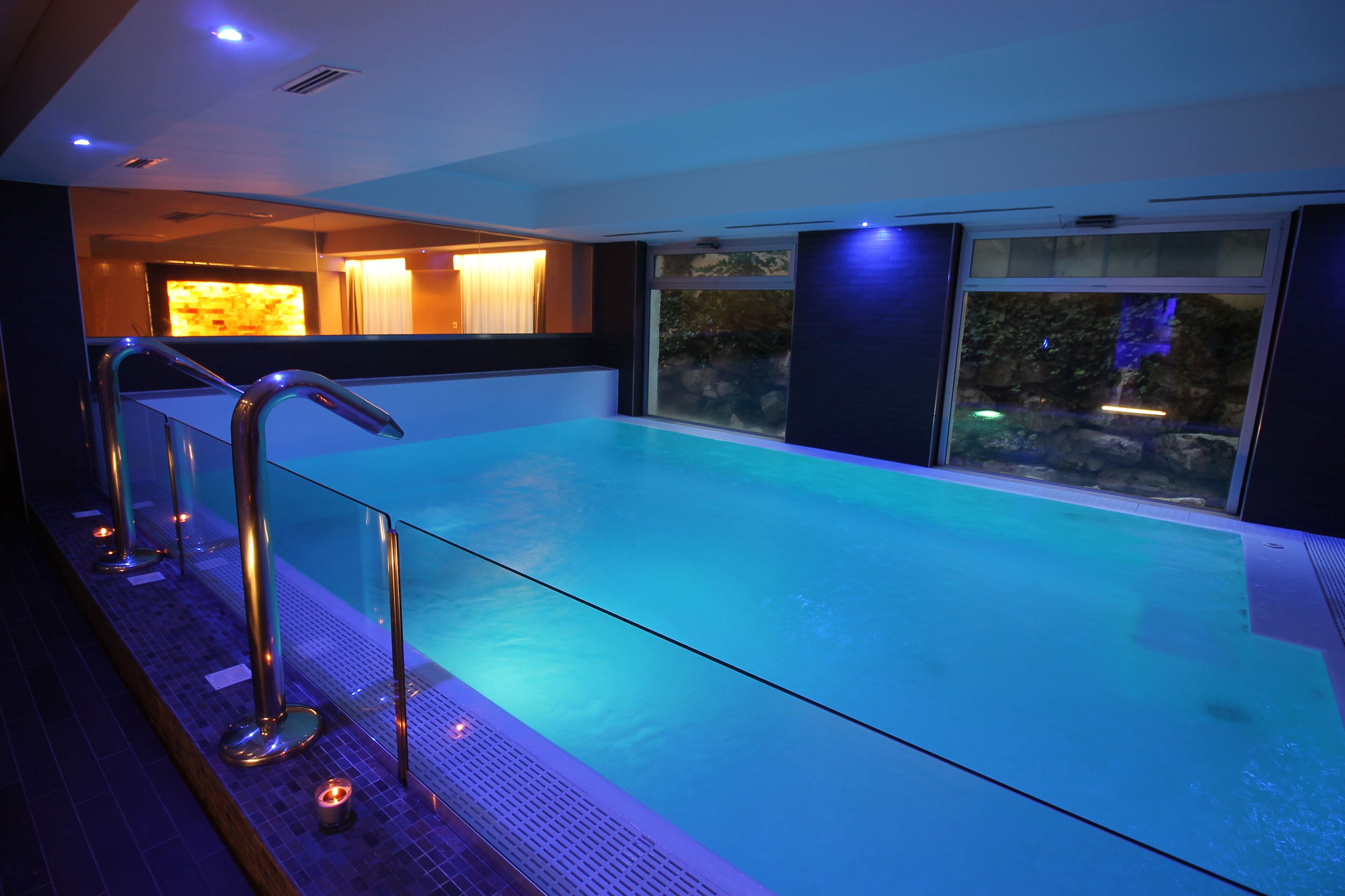 A modern indoor swimming pool with clear glass walls, a metal railing, and a view of the outdoors.
