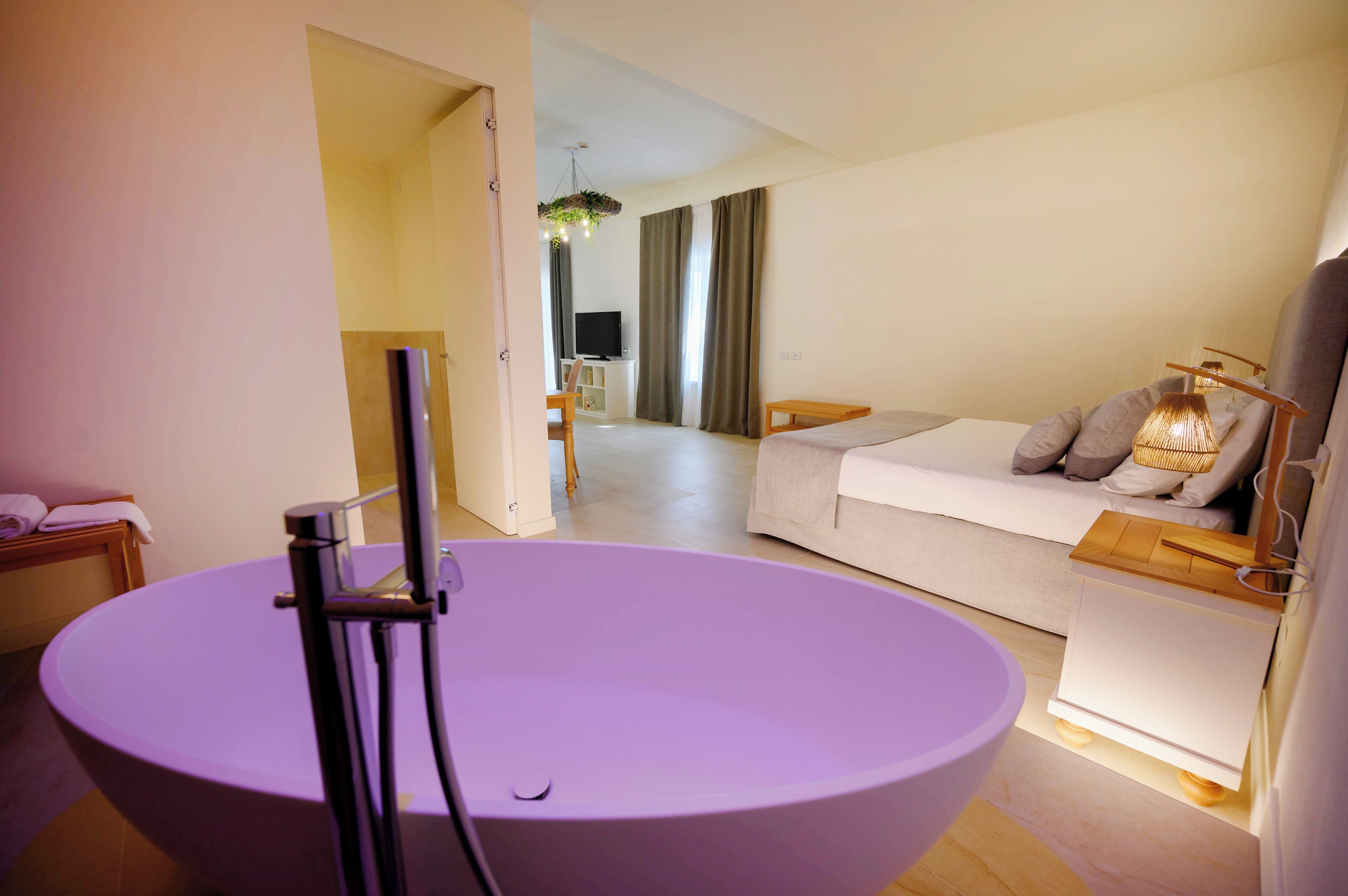 Camera d'albergo moderna con una vasca tonda viola, una divano letto con cuscini, un piccolo tavolino in legno e una televisione sullo sfondo.