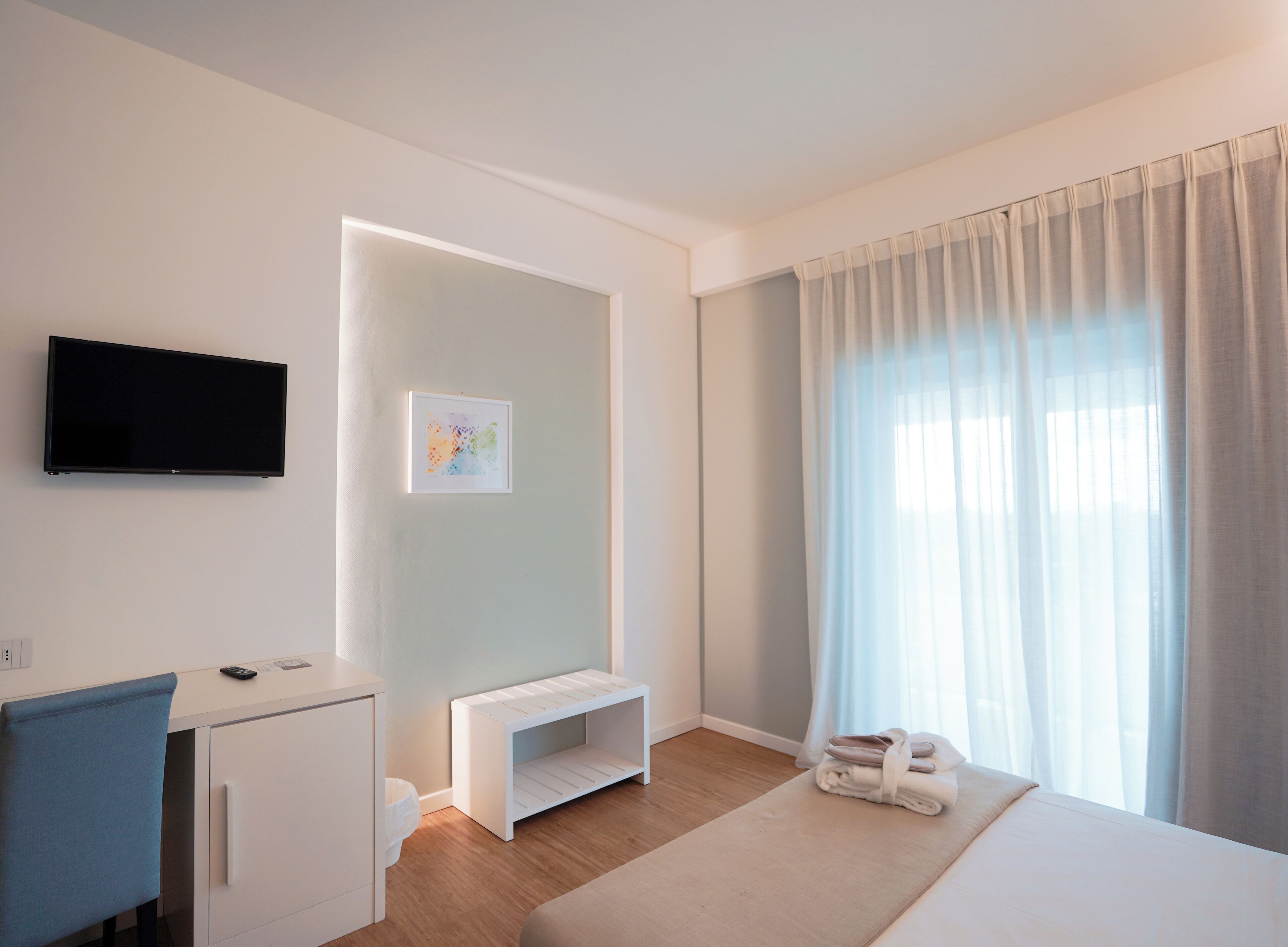 Una habitación de hotel moderna con un televisor de pantalla plana montado en la pared, una pequeña consola blanca, un banco, una cama con cubrecama color beige y un ventanal con cortinas translúcidas.