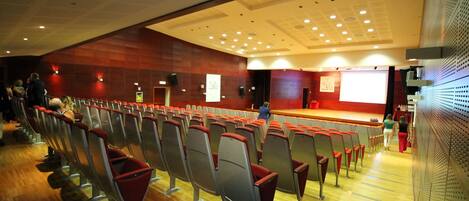Un salón de conferencias con filas de sillas, un escenario y piso de madera.
