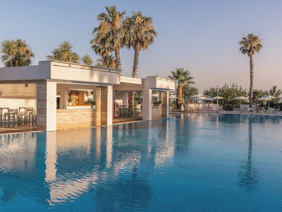 A swimming pool with lounge chairs and umbrellas, surrounded by palm trees and a bar area.