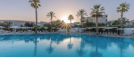 A swimming pool with lounge chairs and umbrellas, surrounded by palm trees, with a building in the background.