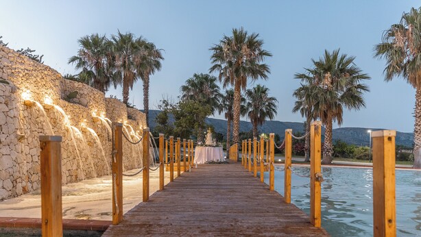 A wooden pathway leading to a pool area with palm trees and a stone wall.