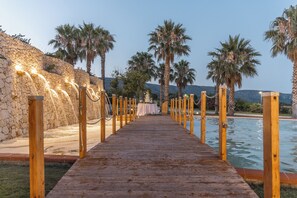 A wooden pathway leading to a pool area with palm trees and a stone wall. - Regiohotel Manfredi (Manfredonia)