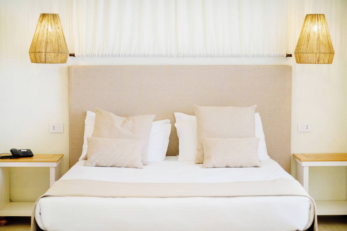 Una cama bien hecha con sábanas y almohadas blancas, dos mesitas de noche con lámparas, y un teléfono sobre la mesa izquierda.