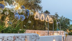A stone wall with a decorative metal structure and string lights, surrounded by lush greenery and flowers.