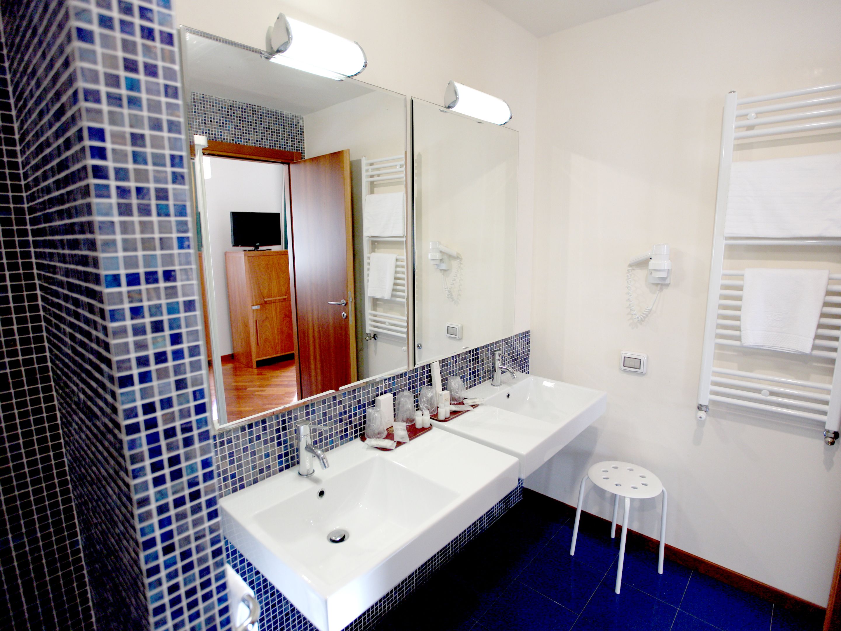 Une salle de bain avec un grand miroir, deux lavabos, des toilettes et un petit tabouret.